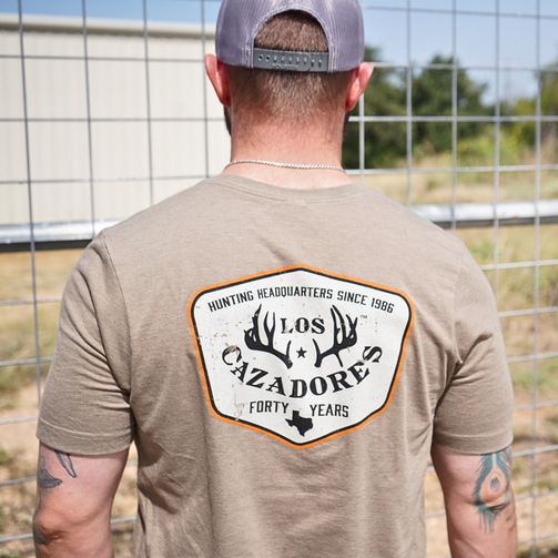 view of the back side of an individual wearing the los cazadores special edition 40th year badge short sleeve tee while standing outside on a ranch.