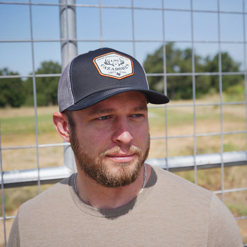 lifestyle image of Los Cazadores trucker cap featuring a black front panel, curved brim, gray mesh back, and commemorative “Forty Years” front patch being worn by a male individual outside in front of a fence looking directly out.