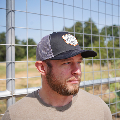 lifestyle image of Los Cazadores trucker cap featuring a black front panel, curved brim, gray mesh back, and commemorative “Forty Years” front patch being worn by a male individual outside in front of a fence lookingto his left.