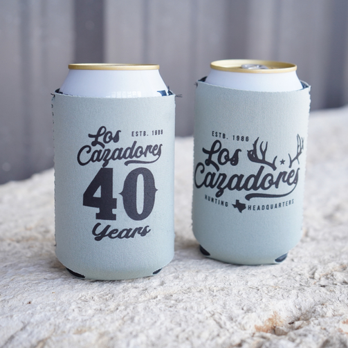 Two Los Cazadores 40 Years commemorative sage green neoprene can cooler holding 12 oz can beverages facing different directions to show the different graphic designs