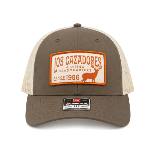 Front view of a Los Cazadores trucker hat with taupe front panels, cream mesh back, orange patch, and curved brim on a white background.