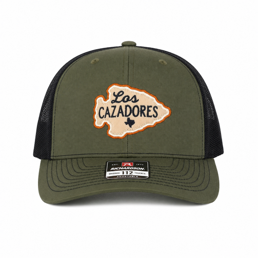 Front view of the Los Cazadores olive and black trucker cap with arrowhead patch and brim sticker on a clean white background.