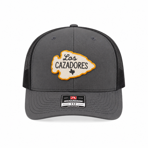 Front view of the Los Cazadores grey and black trucker cap with arrowhead patch and brim sticker on a clean white background.