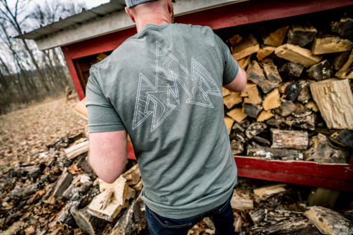 Man wearing the Pnuma Topo Logo Tee Shirt in Green stacking firewood, back view