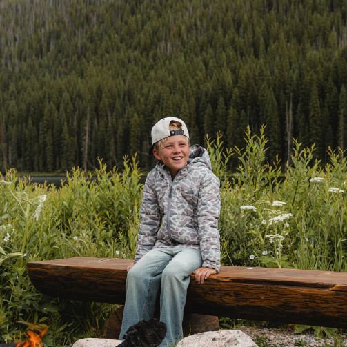 Boy wearing the Burlebo youth puffer jacket in classic deer camo sitting on a bench outdoors