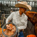 Cowboy in a felt hat holding a lariat beside his horse in a barn while wearing a white check Poncho Western long sleeve pearl snap shirt