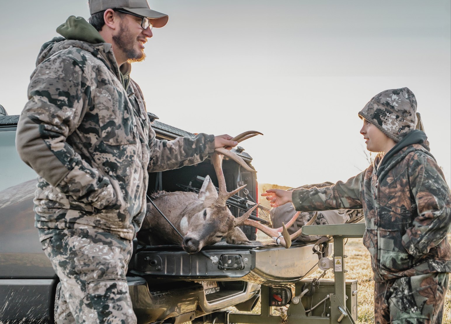 A hunter in camo holds the antlers of a whitetail buck on his truck tailgate while a young hunter reaches out to touch it