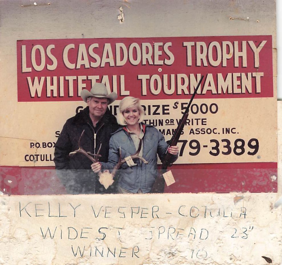 Two hunters pose with a buck in front of the original red and white Los Casadores Trophy Whitetail Tournament sign advertising a $5,000 grand prize, with a handwritten note reading Kelly Vesper, Cotulla, widest spread 23 inch winner