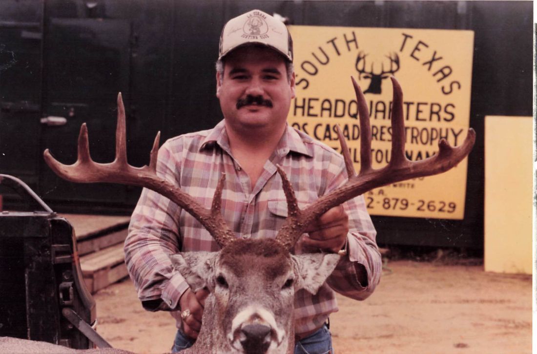 A hunter holds up a heavy-antlered whitetail buck in front of the yellow South Texas Headquarters, Los Cazadores Trophy Whitetail Tournament sign