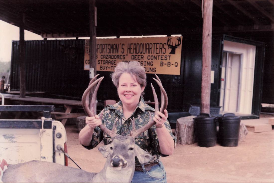 A smiling woman holds up the antlers of her buck beneath a Sportsman's Headquarters, Los Cazadores Deer Contest sign