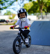 Young boy rides a black Strider 12 Pro bike with custom Carbon Fiber Wheels, Carbon Fiber Flat Handlebar, and yellow Strider Mini-Grips down a black ramp. He's wearing a white full face helmet, a pair of red racing gloves, and a big smile!