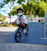 Young boy rides down a black bike ramp on a custom black Strider 12 Pro bike with Carbon Fiber Wheels, Carbon Fiber Flat Handlebar, and yellow grips. He's also wearing a protective full face helmet!