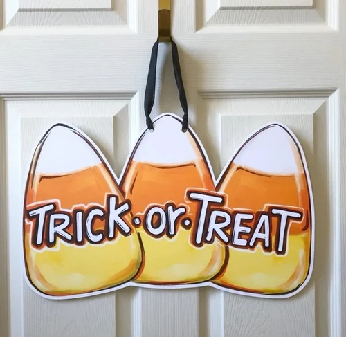 Candy Corn Door Hanger. Featuring a painterly illustration of three candy corn pieces next to each other with "TRICK OR TREAT" overlayed in white text, this design is printed on a white PVC sign, hanging by a black ribbon, waterproof and perfect for indoor or outdoor use. Measures 12in x 20in without ribbon, .25in thick material.