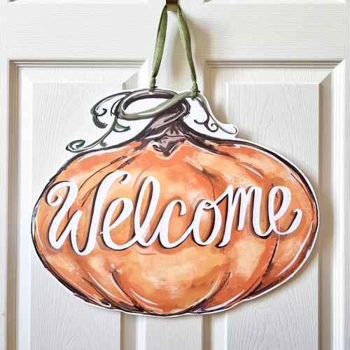 Pumpkin Welcome Door Hanger. Featuring a painterly illustration of an orange pumpkin with a curly brown stem and "Welcome" in white script, this design is printed on a white PVC sign, hanging by a muted green ribbon, waterproof and perfect for indoor or outdoor use. Measures 20in x 16in without ribbon, .25in thick material.
