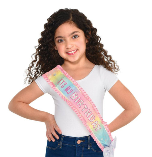 Child Pastel Party Birthday Sash. This shimmery pastel rainbow sash features a pink ribbon trim with iridescent sequins, a large white iridescent bow at the bottom, and "IT'S MY BIRTHDAY" in pink and iridescent text. Measures 4in x 28.5in. One size fits most children.