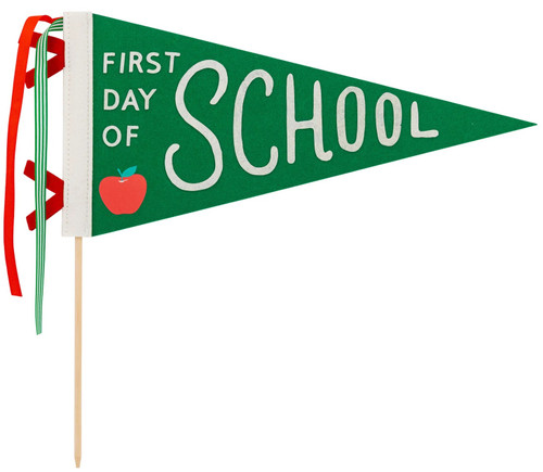 First Day Of School Felt Pennant. This green felt flag pennant features "FIRST DAY OF" in small white letters beside "SCHOOL" in larger white letters, accented with a red apple. It includes red and green ties and a wooden dowel. The underside of the pennant is solid green. Dowel measures 14in, felt pennant measures 15in.