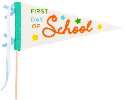 First Day Of School Multicolor Felt Pennant. This off white felt flag pennant features "FIRST DAY OF" in small green, blue, and orange letters beside "SCHOOL" in larger orange script, accented with yellow, blue, and green stars. It includes light blue and white ribbon ties and a wooden dowel. The underside of the pennant is solid off white. Dowel measures 14in, felt pennant measures 15in.