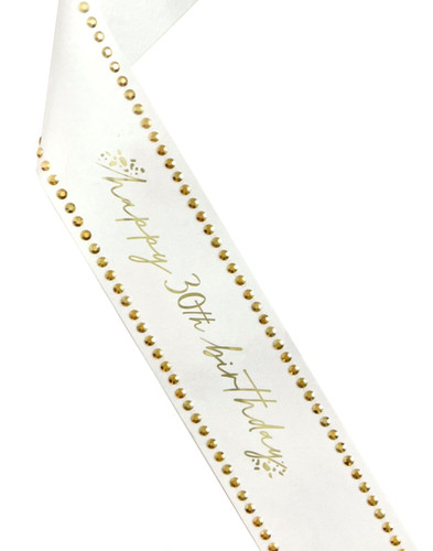 Paper 30th Birthday Sash with Gold Rhinestones. This white cardstock paper sash features "happy 30th birthday" in gold foil script, accented by cluster of gold foil spots framing the text and gold acrylic rhinestones along the edges. One size fits most.