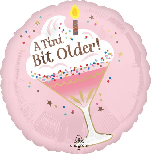 This A Tini Bit Older Birthday Balloon is circular and light pink with a darker pink martini glass topped with white frosting, colorful sprinkles, and a candle, accented with shimmery gold and tilted diagonally. "A Tini Bit Older!" in rose gold text overlays the frosting. Balloon is approximately 17 inches after inflation.