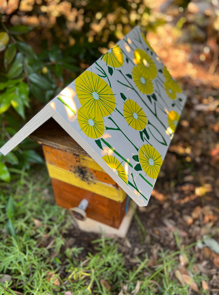 Garden beehive with a white roof hand-painted with bright yellow flowers and green stems.