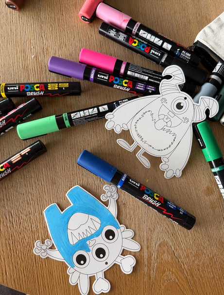POSCA Colour In Monster Cards being coloured with POSCA paint markers, showing partially coloured monster illustrations and assorted markers on a tabletop.