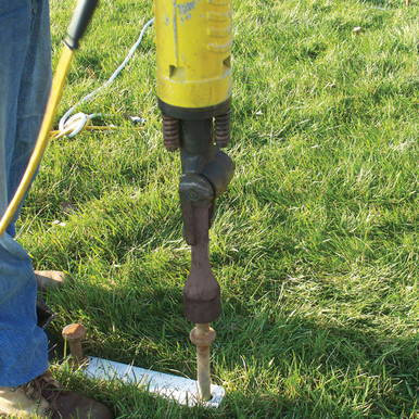 2'' Stake Driver Bit, a close-up photo of a 2-inch drive bit attached to a yellow post driver tool on a grassy surface for driving stakes into the ground.