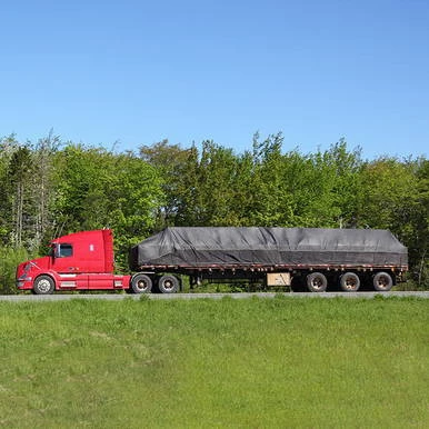 16' x 24' Steel Tarp, 2 Flap, covering a large flatbed truck in a grassy field with trees and a clear blue sky in the background.