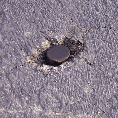 1 1/8'' Asphalt Plug, close-up view showing a black rubber asphalt patch plug embedded in gray asphalt surface.