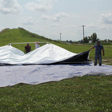 25' x 25' Polyester Drop Cloth, people setting up the large white and black drop cloth on grass outdoors with a hill and power lines in the background.