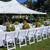 Oxford White Resin Chair, outdoor wedding or event seating arrangement with white resin chairs arranged around a long banquet table with floral centerpieces, under large white event tents on a grassy lawn.
