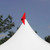 Red Pole Flag, a vivid red flag attached to the top of a white tent peak against a clear blue sky with trees in the background.