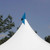Blue Pole Flag, a vibrant blue flag attached to a white pole at the top of a tent during daytime with trees and a clear sky in the background.
