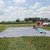 25' x 25' Polyester Drop Cloth, outdoor scene with the large protective cloth spread out on grass, people setting up the drop cloth near a car under a partly cloudy sky.