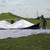 35' x 35' Polyester Drop Cloth, image shows workers setting up or adjusting a large white drop cloth outdoors on grassy terrain with a hill and power lines in the background.