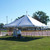 Celina Tent 20' x 20' Classic Series Pole Tent, 1 Piece Tent Top, Complete - A large white event tent with a peaked roof set up outdoors on a grassy area with support poles and cords, under a clear blue sky.