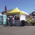 Celina Tent 10' x 10' Pinnacle Series High Peak Frame Tent / Cross Cable Marquee, complete setup at outdoor event with people gathered around and a yellow and white canopy.