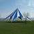 52' TP Series Hexagon Tent, Complete, outdoor event tent with blue and white striped canopy and supporting stakes on a grassy area near a body of water.