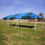 Celina Tent 10' x 30' Classic Series Frame Tent, 1 Piece Tent Top, Complete, outdoor event tent with blue and green canopy setup on a grassy field under a clear sky.