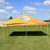 Celina Tent 20' x 20' Classic Series Frame Tent Full Digital Print, complete outdoor event tent with a bright yellow and orange canopy, featuring a blue and white striped logo, set up on a grassy field under a partly cloudy sky.