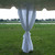 9' x 40" Leg Drape with Sash, white fabric draped and tied at the bottom, outdoors on a grassy field with water and trees in the background.
