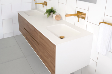 Wood-style vanity with white countertop, double sinks, taps, and a green plant against a white wall, with a gold towel rail.