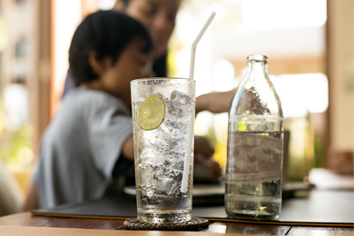 Filtered water served in a glass bottle alongside glasses of water with lemon slices and straws, with people in the background