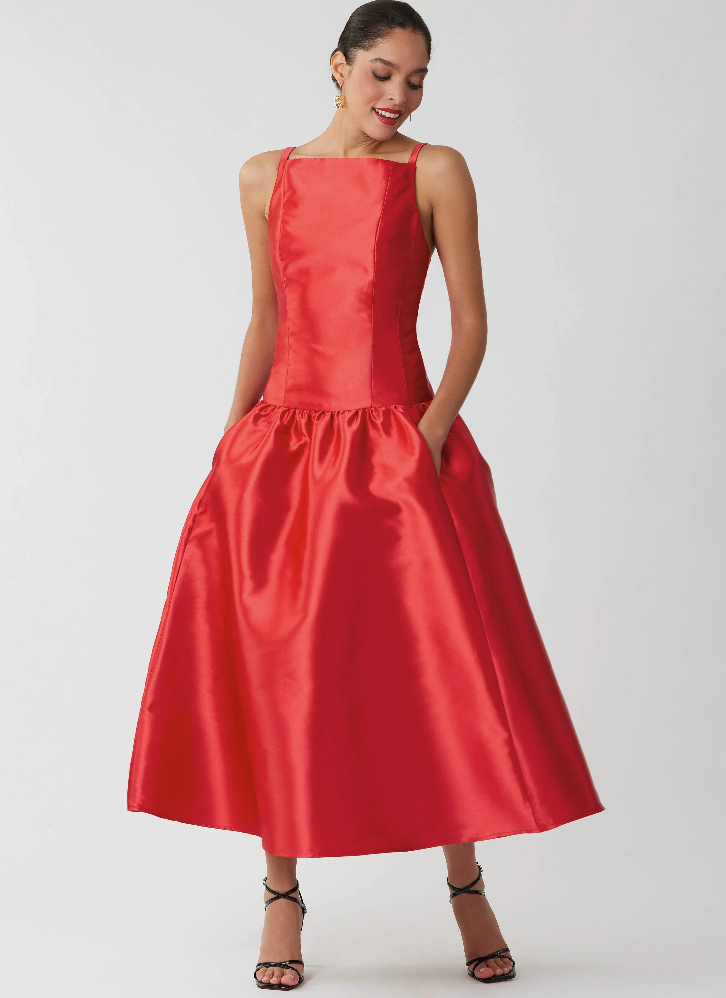 Model wearing a red satin tea length dress with black strappy sandals.