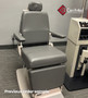 Reliance 6200H ENT exam chair in a clinical room, shown in an upright position with gray upholstery, square adjustable headrest, flat padded armrests, and rectangular base, placed next to medical equipment cabinet.