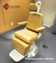 Reliance 980 ENT procedure chair with yellow upholstery, adjustable headrest, armrests, and footrest, placed on a silver circular base in a clinical room with gray flooring and tan walls.