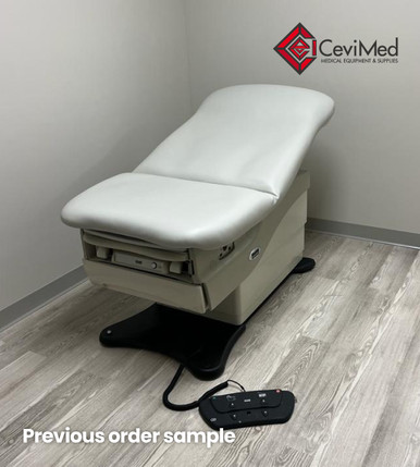 Midmark 625 bariatric treatment table with gray upholstery shown in a medical exam room, positioned against a white wall and sitting on light wood flooring, with the foot control visible on the floor.