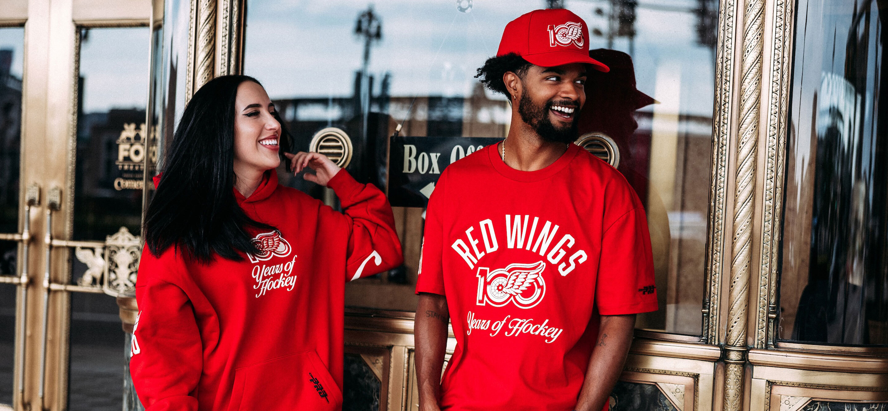 Man and woman standing outside Fox Theatre wearing 100 years of Hockeytown apparel. Comerica park can be seen in the reflection of the windows of the Fox Theatre. data-size=