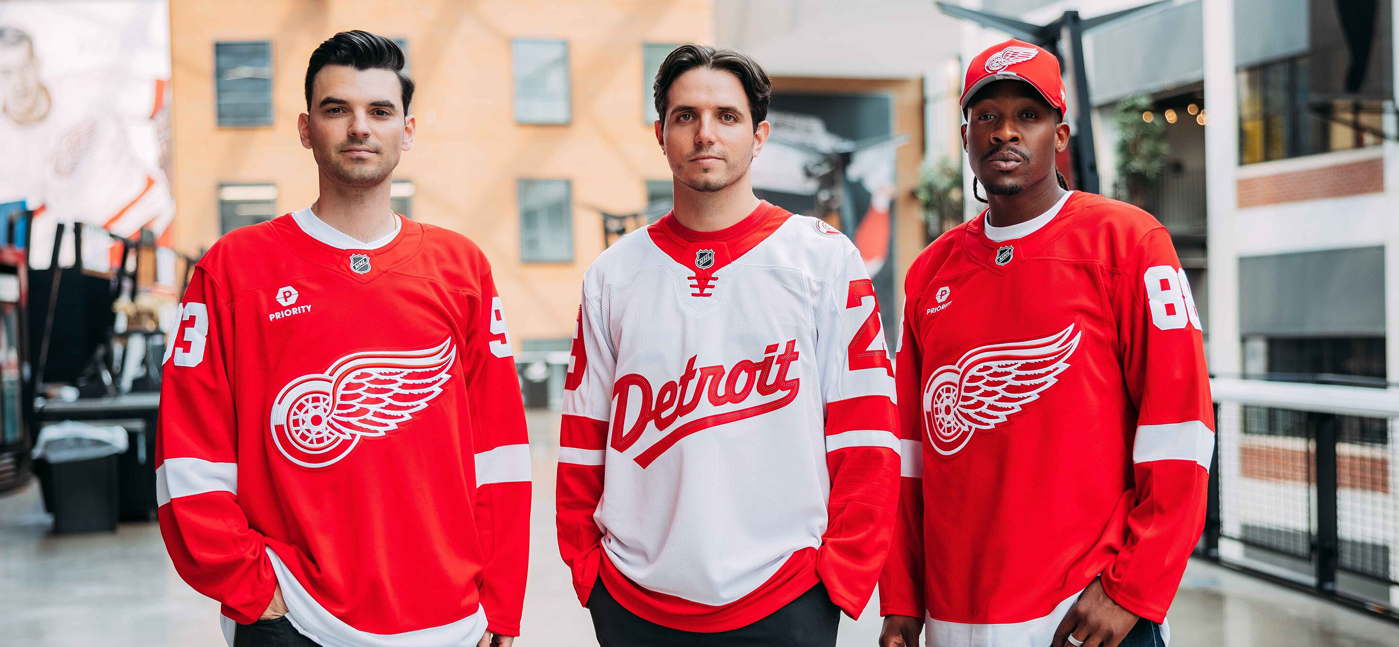 Three men posing inside of the hockey arena wearing red and white stadium series Detroit Red Wings jerseys with the logo on the front. data-size=