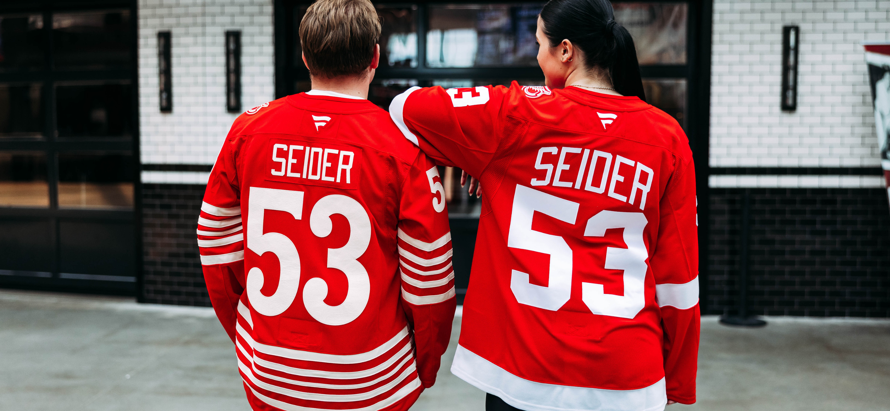 Male and female modeling the Detroit Red Wings Seider Home Premium jersey and Red Wings Seider Centennial Jersey data-size=