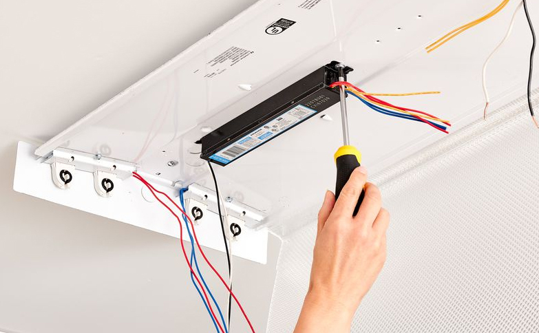 Person using a screwdriver to install or replace an electronic ballast in a fluorescent light fixture mounted on the ceiling.